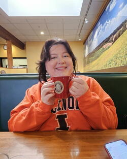 picture of Natalie Wyatt sitting in a booth wearing an orange Illinois crewneck, holding a mug and smiling.